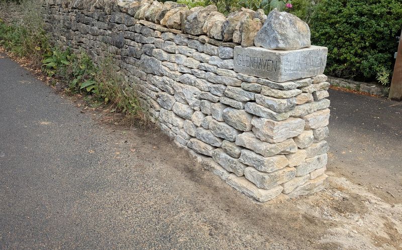 Rebuilt Cotswold stone wall and pier beside a lane, with hollyhocks